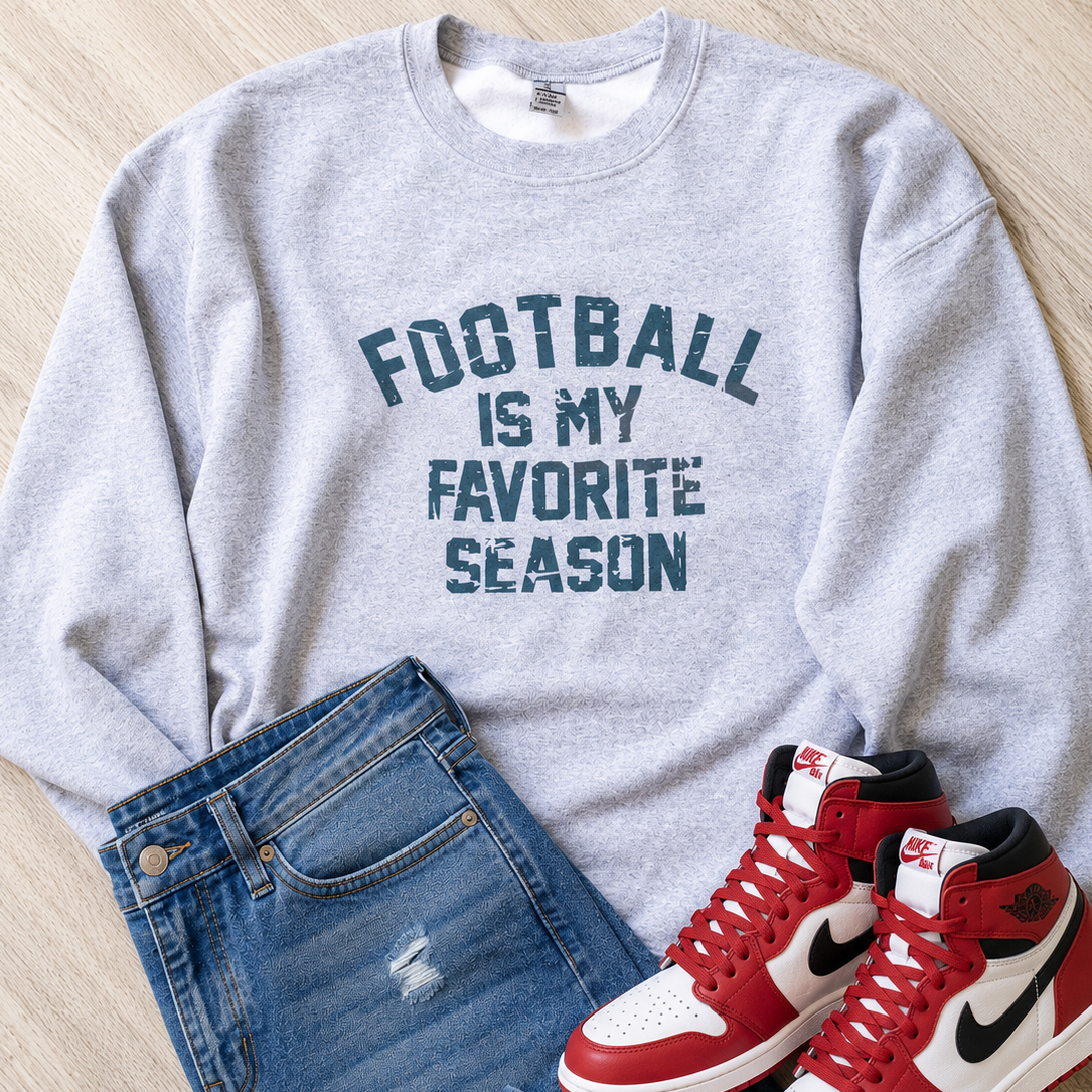 A light heather gray crewneck sweatshirt with the text 'FOOTBALL IS MY FAVORITE SEASON' in dark blue, styled in a flat lay with blue jeans and red and white high-top sneakers on a light wood floor.