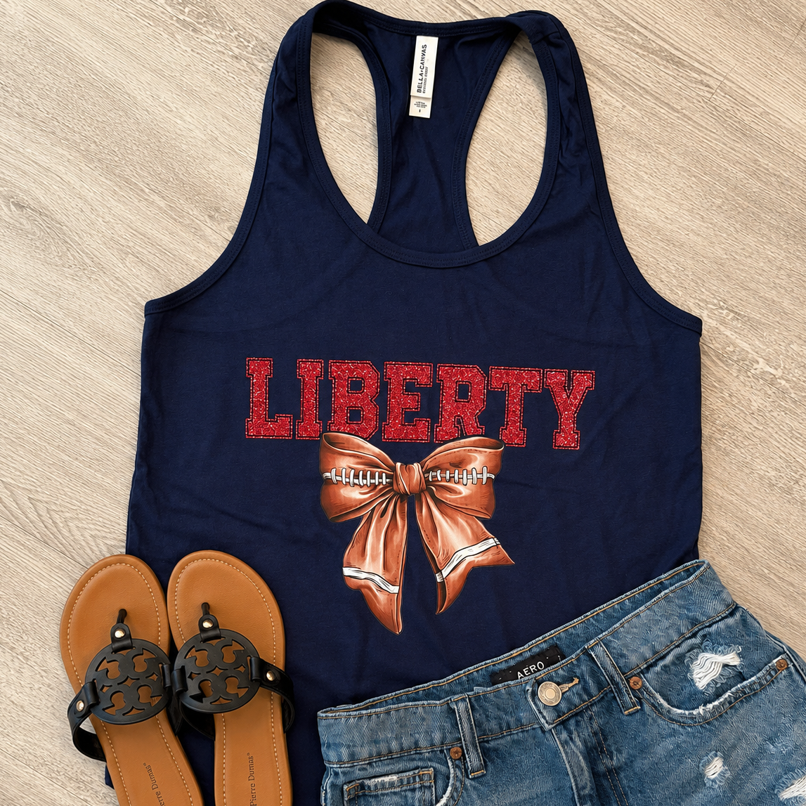 A navy blue racerback tank top with a red 'LIBERTY' text graphic and a brown football-themed bow design, styled in a flat lay with blue denim shorts and brown sandals on a light wood floor.