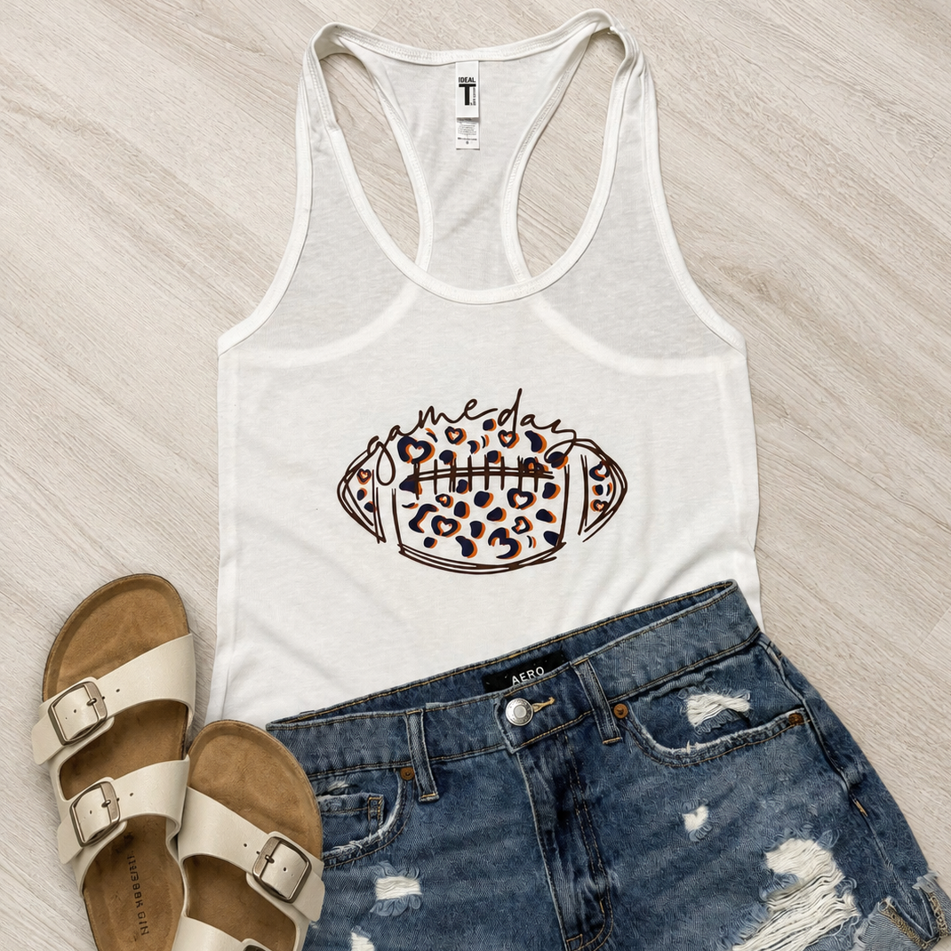 A white racerback tank top with a leopard-print football graphic, styled in a flat lay with distressed denim shorts and beige slide sandals on a light wood floor.