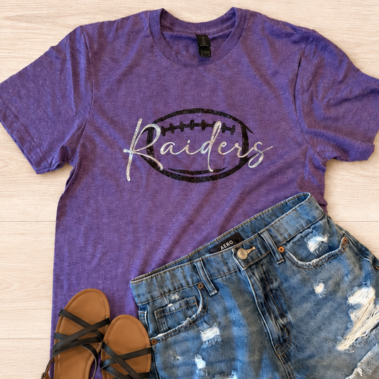 A purple heather graphic t-shirt with 'Raiders' in white script over a black football outline, styled in a flat lay with distressed denim shorts and black sandals on a light wood background.