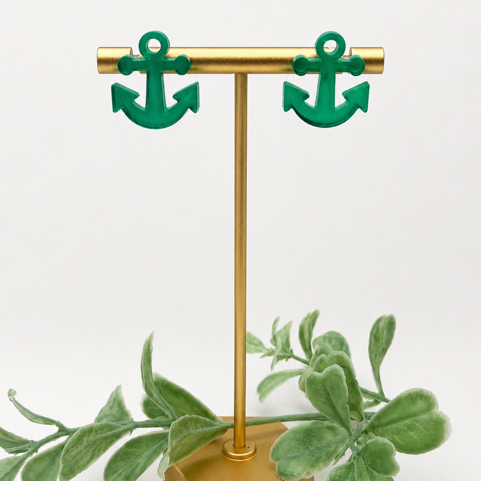 A pair of green anchor-shaped stud earrings displayed on a gold T-stand with a gold hexagonal base, styled with green leaves against a white background.