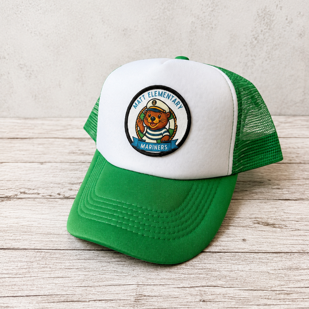 A green and white trucker hat with a circular 'Matt Elementary Mariners' patch on the front, displayed on a light wood surface against a textured light gray wall.