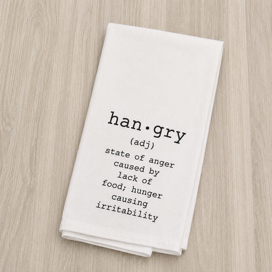 A folded white kitchen towel with black printed text defining 'han·gry' as anger caused by lack of food, hunger, and irritability, displayed on a light wood background.