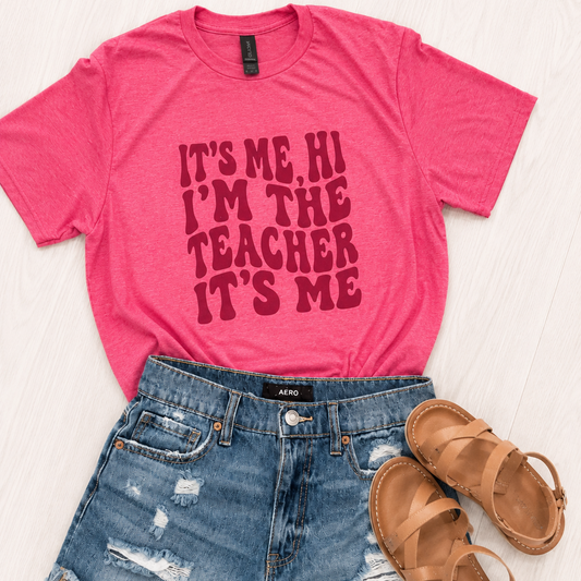 A pink graphic t-shirt with the text 'IT'S ME, HI I'M THE TEACHER IT'S ME' in dark red, styled in a flat lay with distressed blue denim shorts and tan leather sandals on a light wood background.