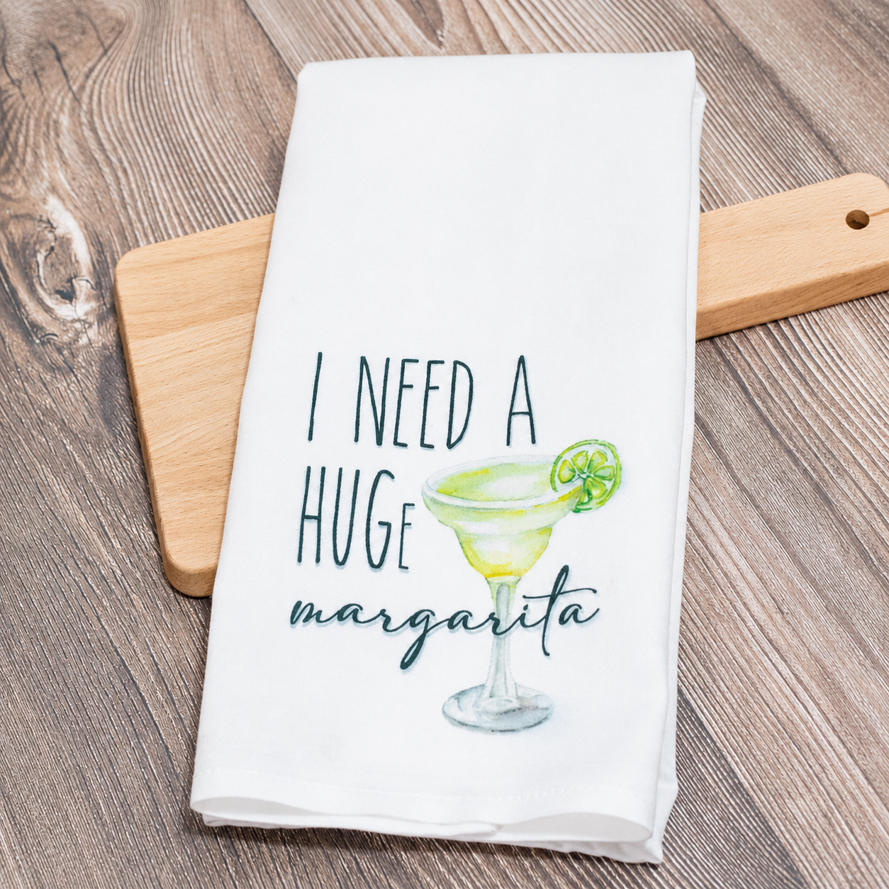A white kitchen tea towel with a watercolor-style green margarita glass and the text 'I NEED A HUGE margarita' in dark teal lettering, styled on a wooden cutting board over a wood-grain tabletop.