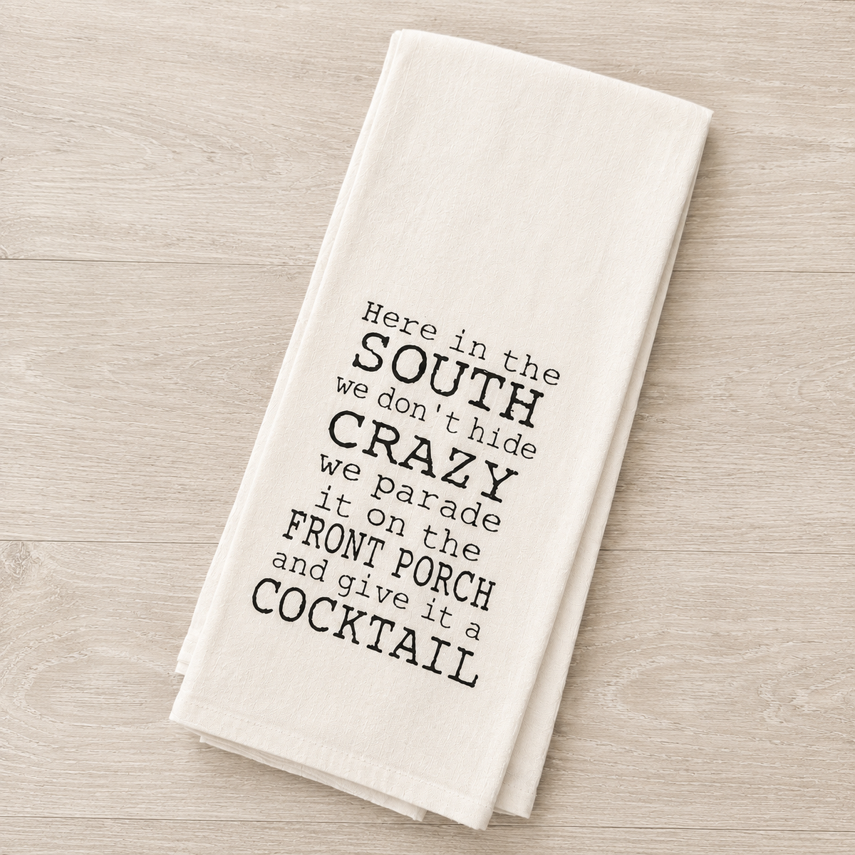 A folded off-white kitchen towel with black printed text reading 'Here in the SOUTH we don't hide CRAZY we parade it on the FRONT PORCH and give it a COCKTAIL', displayed on a light wood surface.
