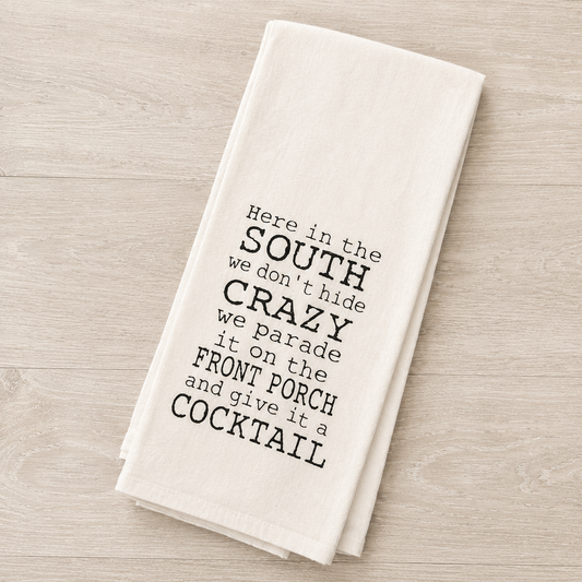 A folded off-white kitchen towel with black printed text reading 'Here in the SOUTH we don't hide CRAZY we parade it on the FRONT PORCH and give it a COCKTAIL', displayed on a light wood surface.