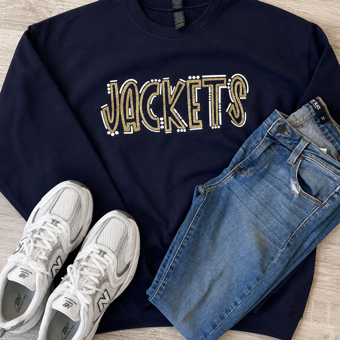 A navy blue crewneck sweatshirt with 'JACKETS' written across the chest in a gold and white outlined collegiate font, styled in a flat lay with blue jeans and white New Balance sneakers on a light wood floor.