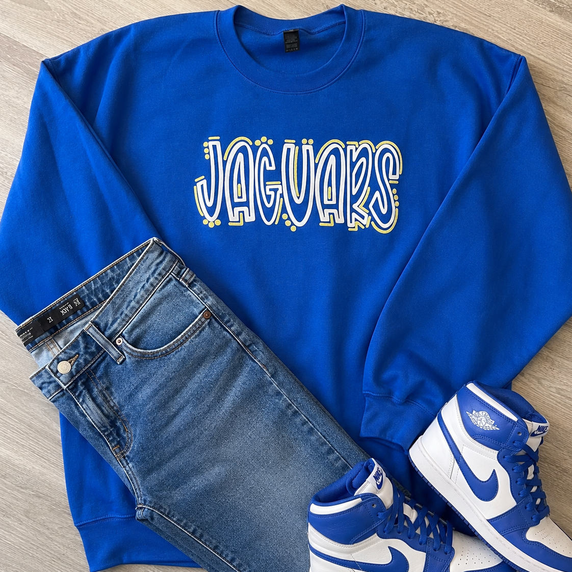 A flat-lay outfit arrangement featuring a royal blue sweatshirt with 'JAGUARS' printed across the chest, paired with blue jeans and blue-and-white high-top sneakers on a light wood floor.