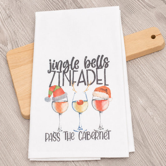 A white kitchen towel with a festive Christmas-themed wine print, styled on a light wood cutting board over a gray wood-grain surface.