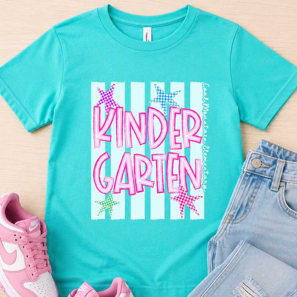 A turquoise short-sleeve graphic T-shirt laid flat on a light wood background, styled with pink sneakers and light blue jeans. The shirt has a large front graphic reading 'KINDER GARTEN' in pink over white vertical stripes with small colorful star shapes.