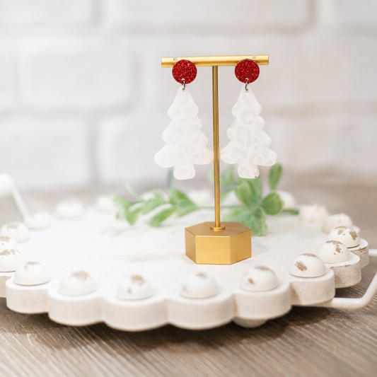 A pair of white Christmas tree-shaped dangle earrings with red glitter studs, displayed on a gold stand over a white ceramic tray.