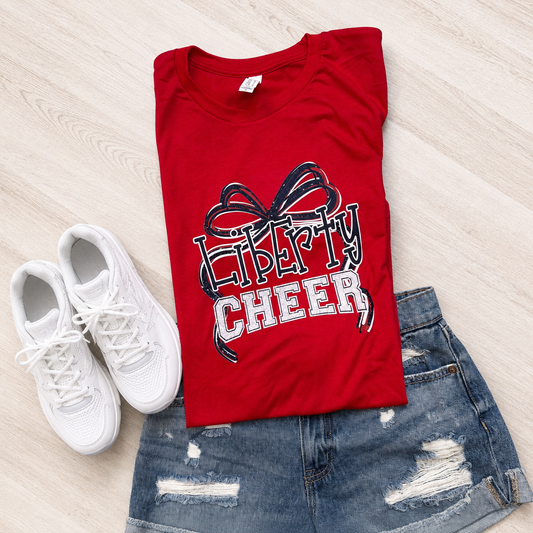 A folded red graphic T-shirt with a black and white 'Liberty Cheer' bow design, styled in a flat lay on a light wood floor with white sneakers and distressed denim shorts.