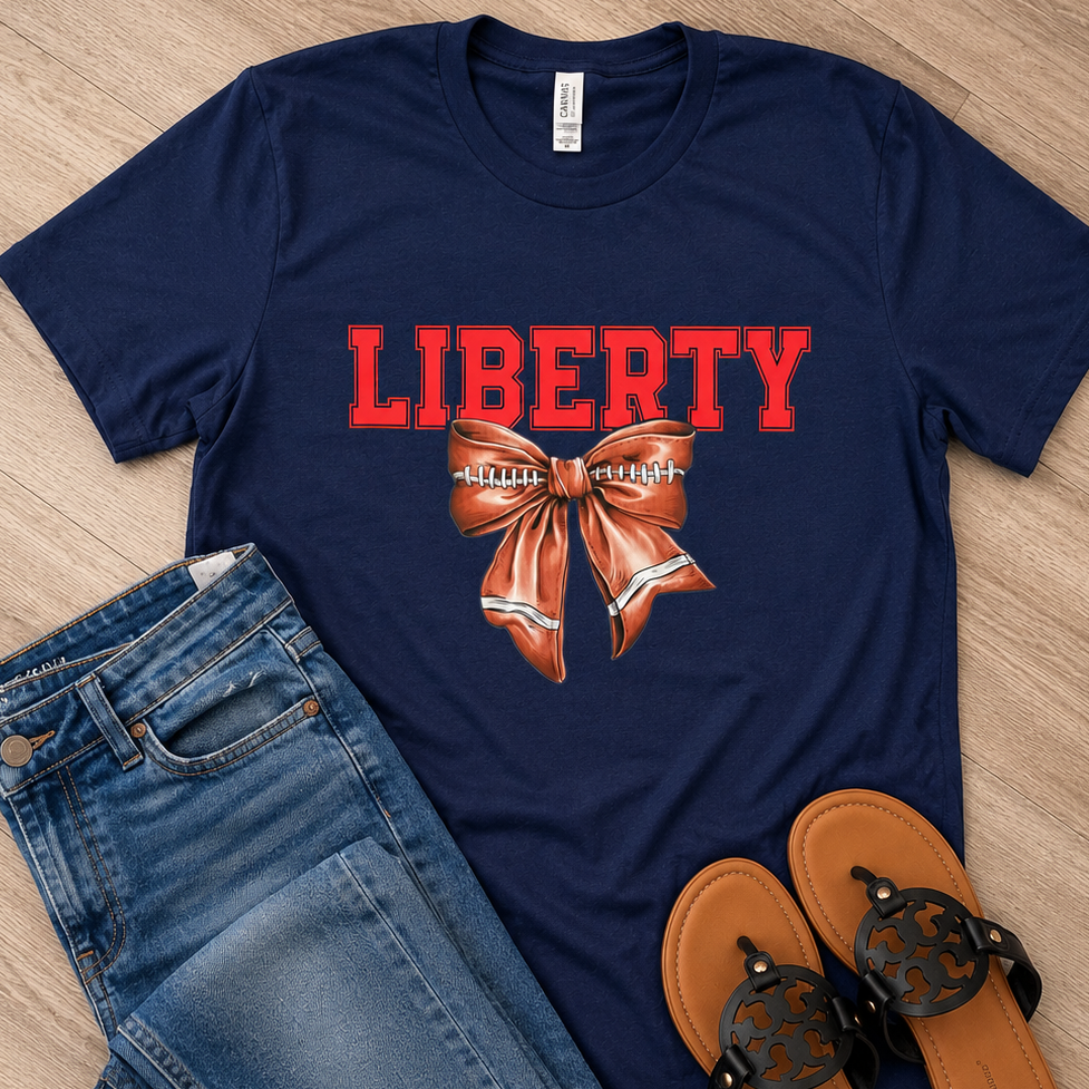 A navy blue graphic t-shirt featuring the word 'LIBERTY' in red and a brown and white football bow graphic, styled in a flat lay with blue jeans and brown sandals on a light wood floor.