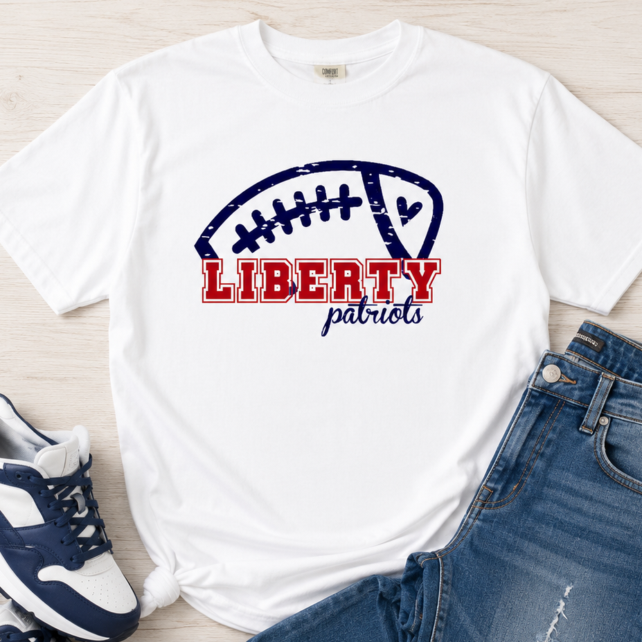 A white graphic T-shirt laid flat on a light wood surface, styled with blue jeans and navy-and-white sneakers. The shirt features a distressed football graphic with the words 'LIBERTY patriots' in red and navy.