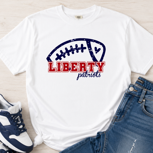A white graphic T-shirt laid flat on a light wood surface, styled with blue jeans and navy-and-white sneakers. The shirt features a distressed football graphic with the words 'LIBERTY patriots' in red and navy.