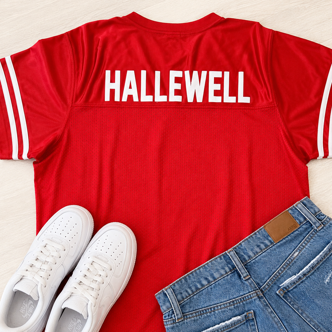 A red short-sleeve athletic-style T-shirt with white stripes on the sleeves and the word 'HALLEWELL' printed in white across the upper back, styled in a flat lay with white sneakers and blue jeans on a light wood background.