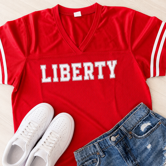 A red short-sleeve V-neck T-shirt with white stripes on the sleeves and 'LIBERTY' printed across the chest in white block letters, styled in a flat lay with white sneakers and distressed blue jeans on a light wood background.