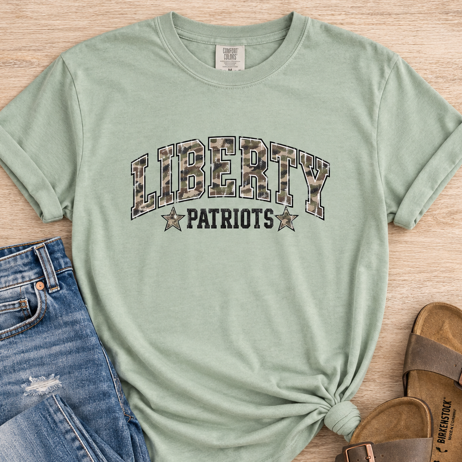 A sage green graphic t-shirt with a knotted hem, styled in a flat lay with blue jeans, brown slide sandals, and a wooden background. The t-shirt features a camouflage patterned 'LIBERTY' graphic and 'PATRIOTS' text with stars.