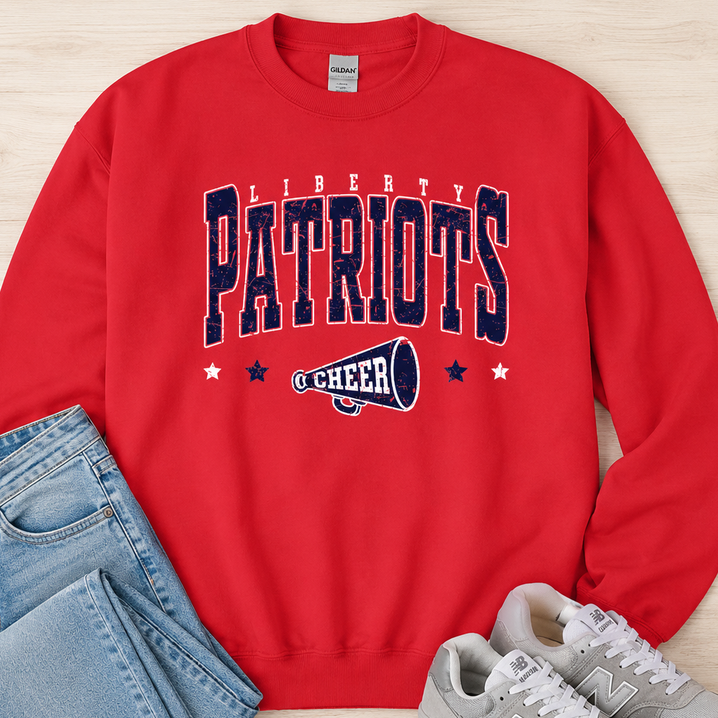 A red crewneck sweatshirt with 'LIBERTY PATRIOTS CHEER' printed in navy and white on the front, styled in a flat lay with blue jeans and gray New Balance sneakers on a light wood background.