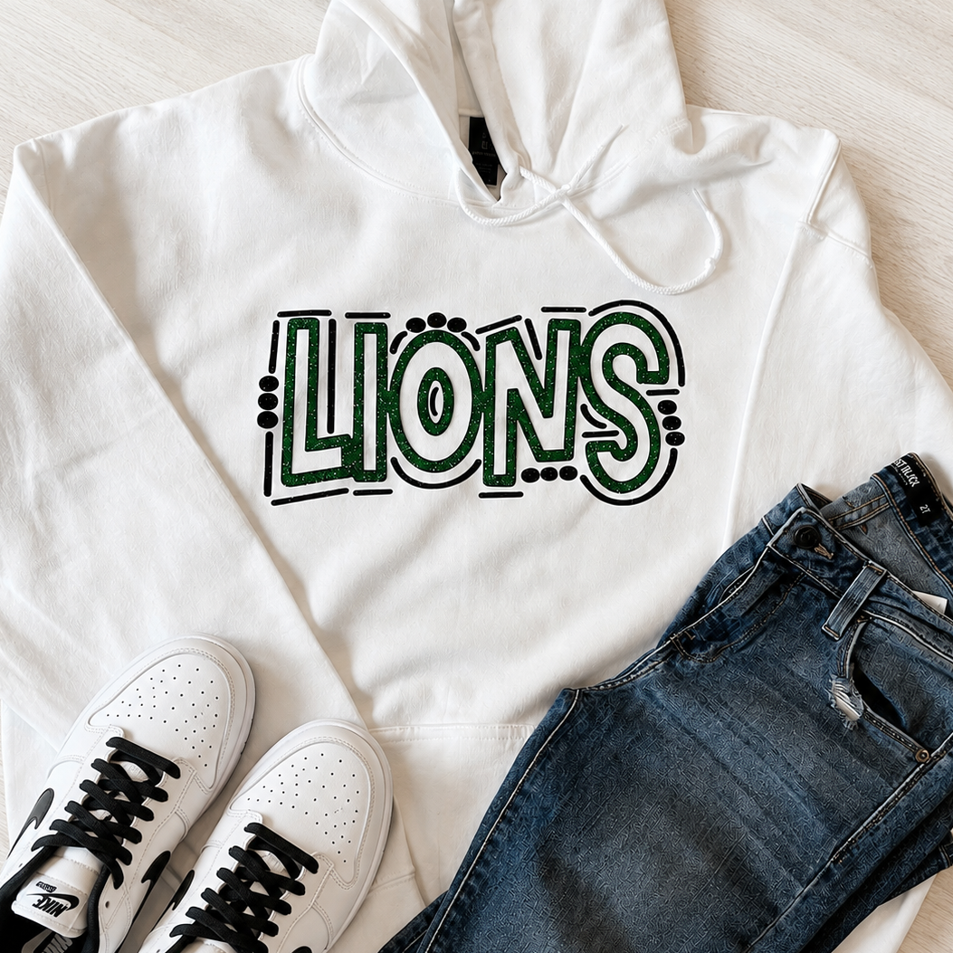 A styled flat-lay featuring a white hoodie with a green and black 'LIONS' graphic, paired with dark blue jeans and white low-top sneakers on a light wood floor.