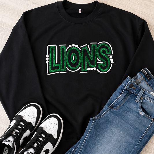 A black crewneck sweatshirt with a green and white glittery 'LIONS' graphic, styled in a flat lay with blue jeans and black-and-white sneakers on a light wood floor.