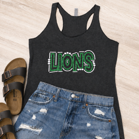A flat-lay outfit featuring a black racerback tank top with a distressed green and white 'LIONS' graphic, paired with light-wash distressed denim shorts and black slide sandals on a light wood background.