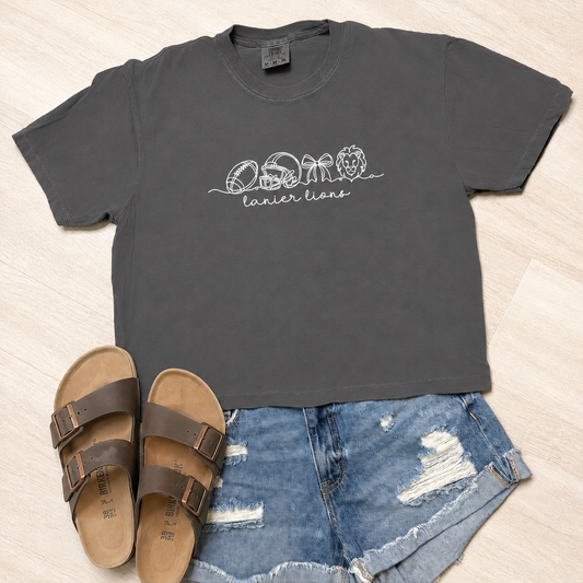 A dark charcoal gray t-shirt with a white graphic of seashells and the words 'osio lanier lions' is styled in a flat lay with blue denim shorts and brown sandals on a light wood floor.