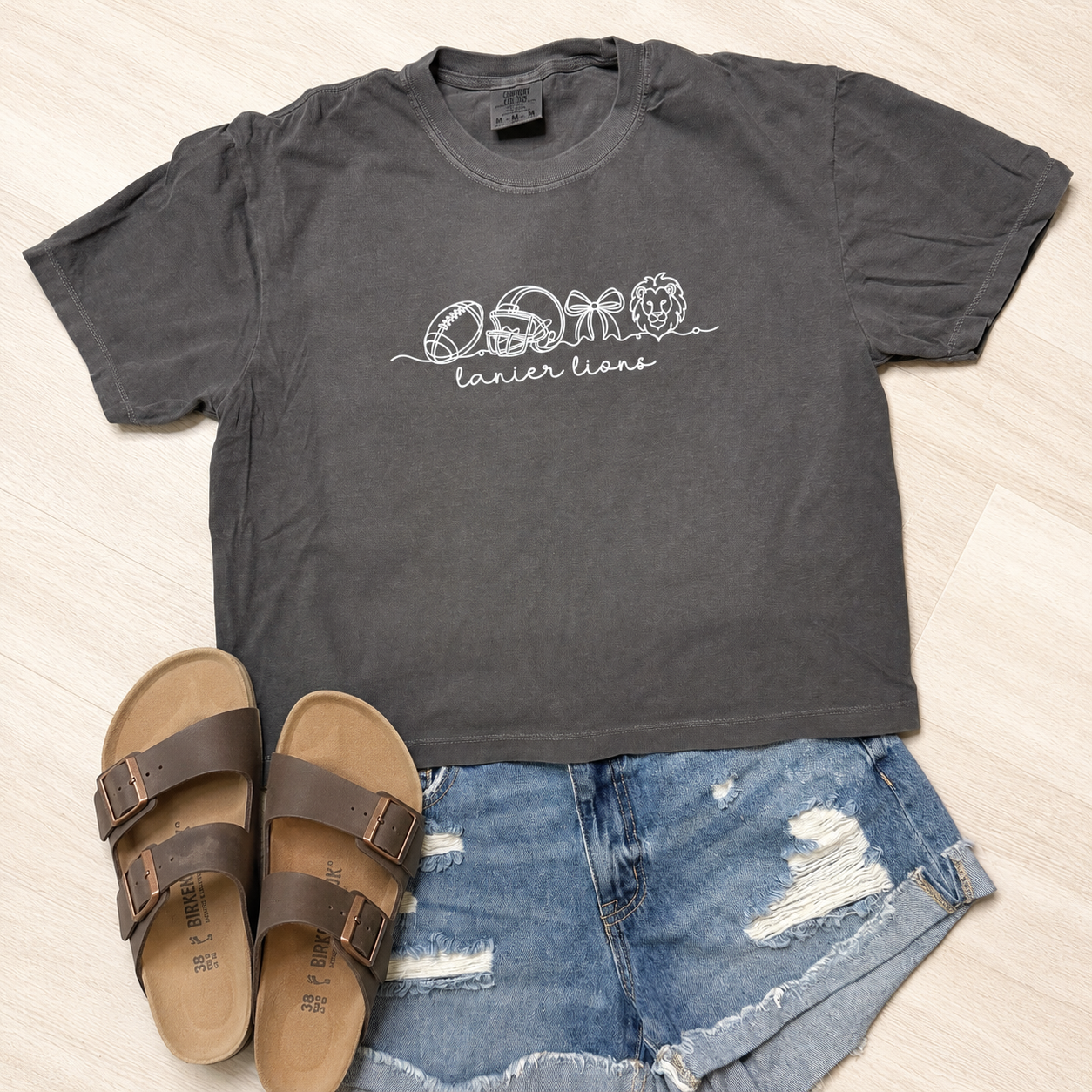 A dark charcoal gray graphic t-shirt with white printed text reading 'OSMO lanier lions' and animal head illustrations, styled in a flat lay with blue denim shorts and brown slide sandals on a light wood floor.