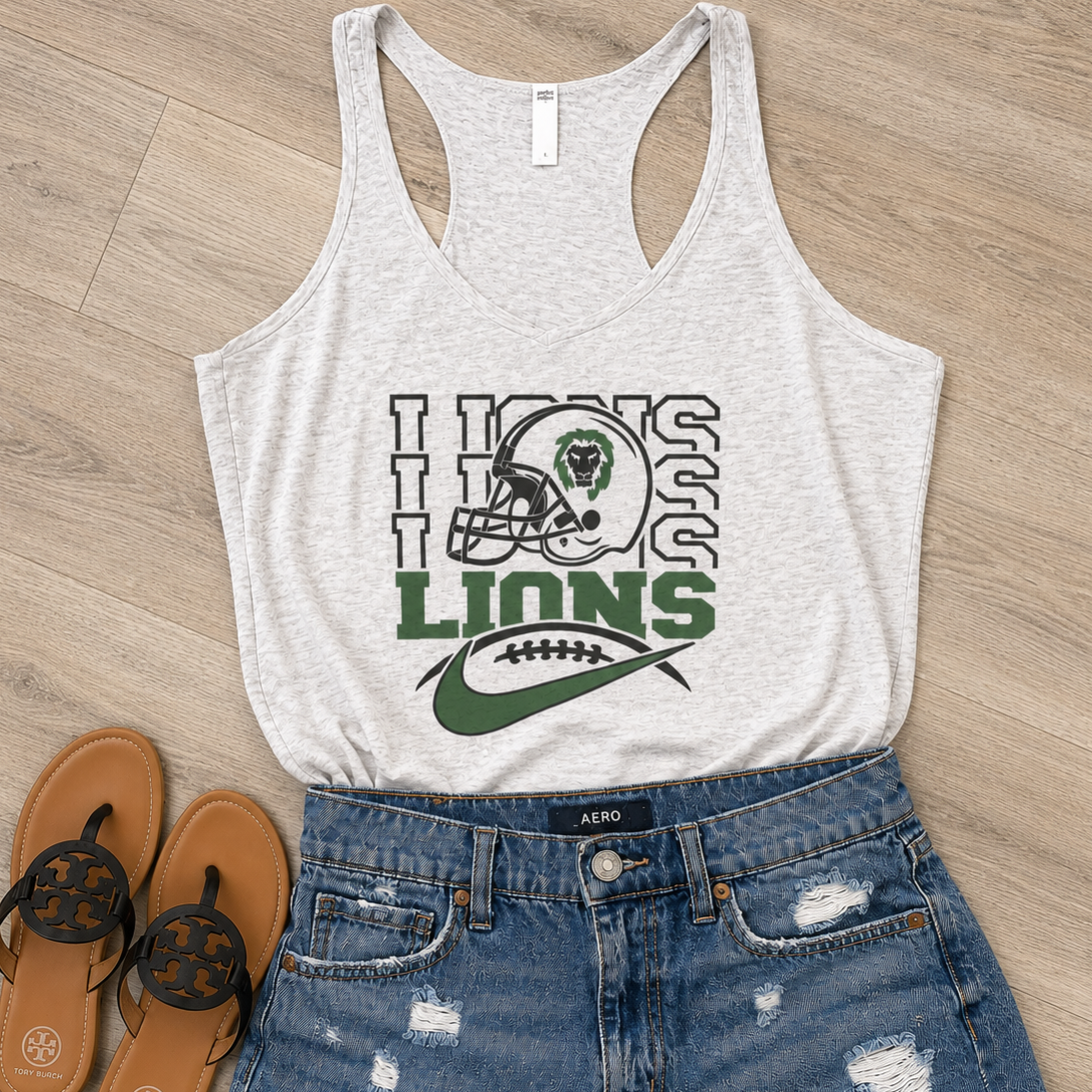 A heather light gray racerback tank top with a green and black 'Lions' football graphic, styled in a flat lay with distressed blue denim shorts and brown flip-flops on a light wood floor.