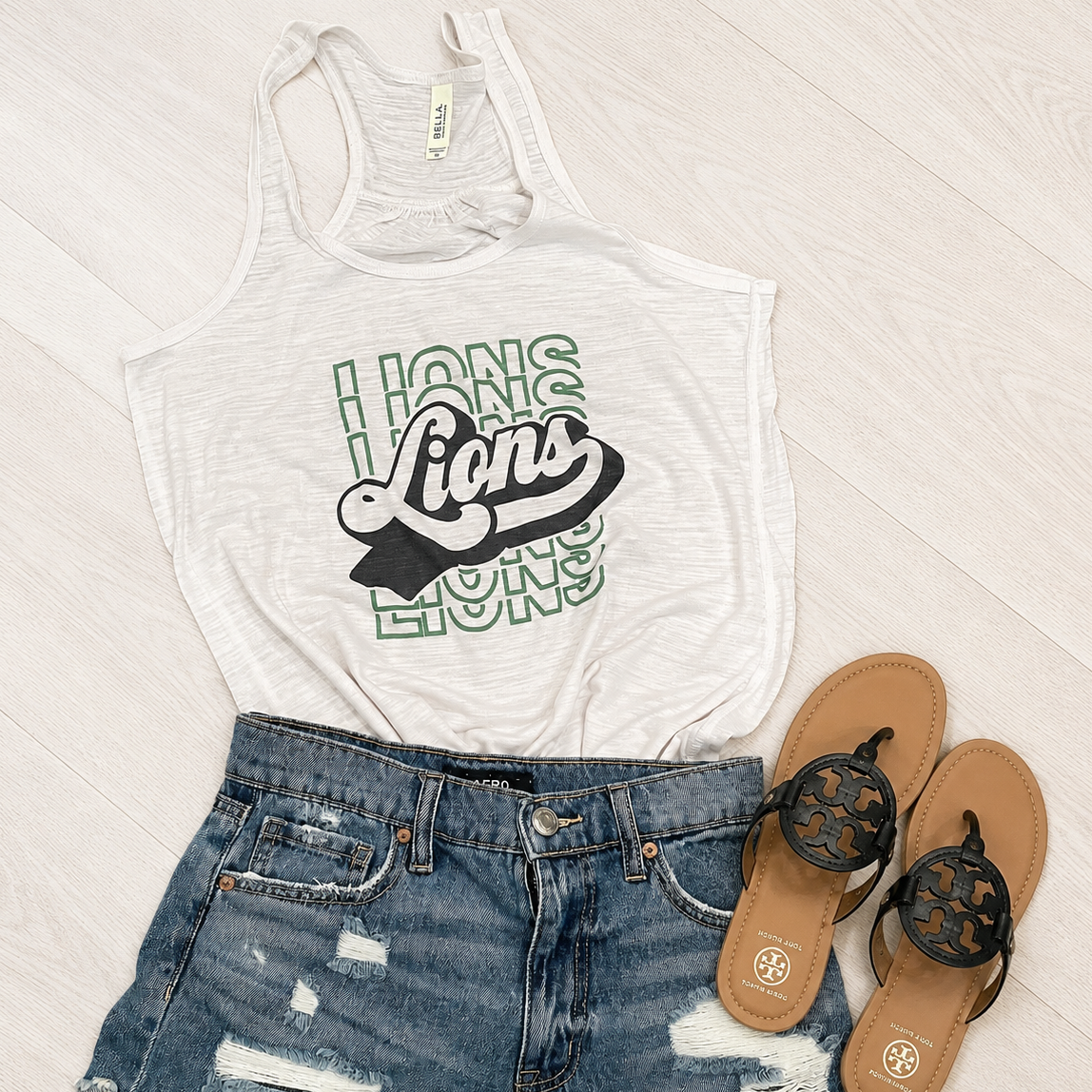 A white racerback tank top with a green and black 'Lions' graphic, styled in a flat lay with distressed blue denim shorts and brown flip-flops on a white wood floor.