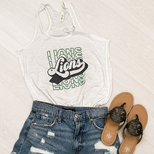 A white racerback tank top with a green and black 'Lions' graphic, styled in a flat lay with distressed blue denim shorts and brown flip-flops on a white wood floor.