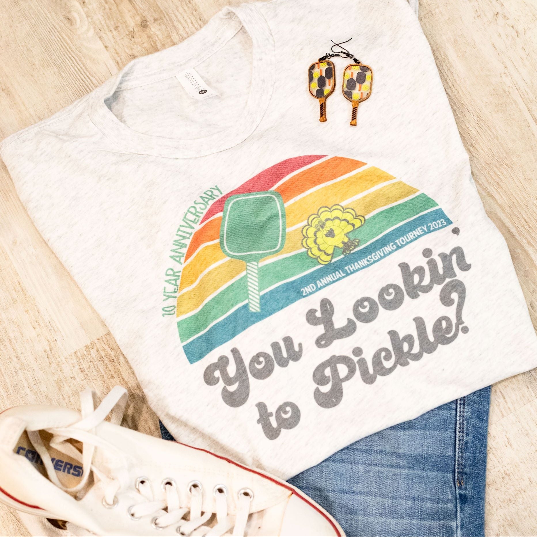 A gray t-shirt with a colorful graphic design and the text 'YOU LOOKIN' TO PICKLE?' on it, displayed flat with a pair of white sneakers and a pair of blue jeans to the side.