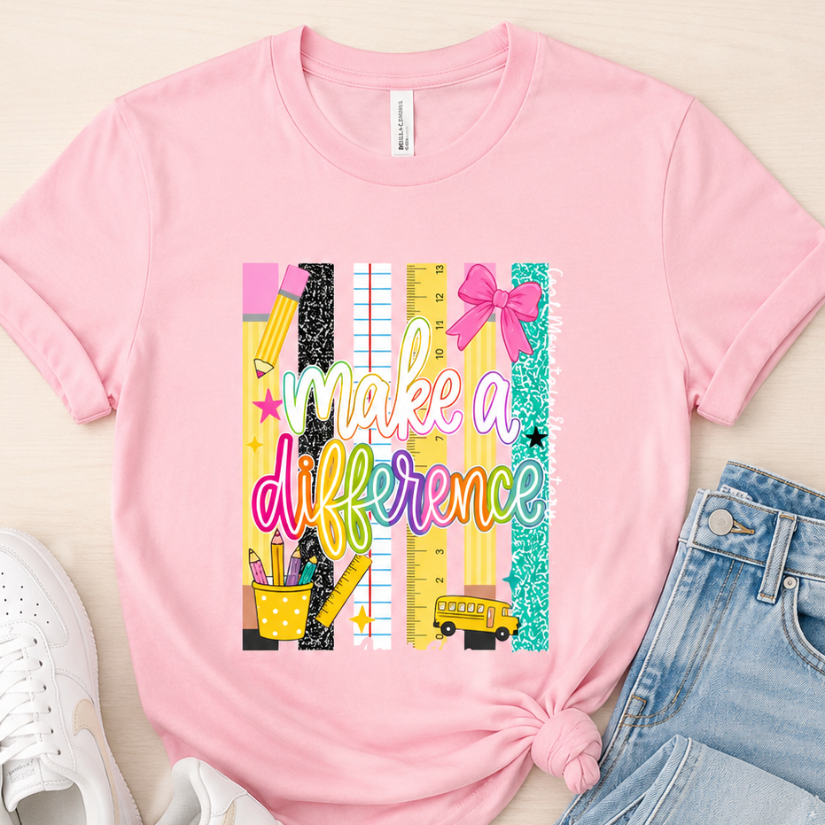 A pink short-sleeve graphic T-shirt laid flat on a light wood background, styled with white sneakers and light blue jeans. The shirt features a colorful school-themed graphic with the phrase 'make a difference' over ruler, pencil, and ribbon illustrations.