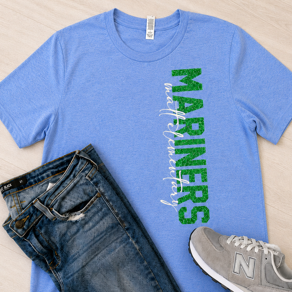 A flat-lay outfit featuring a heather blue t-shirt with a green 'MARINERS' graphic, styled with dark blue jeans and gray New Balance sneakers on a light wood background.