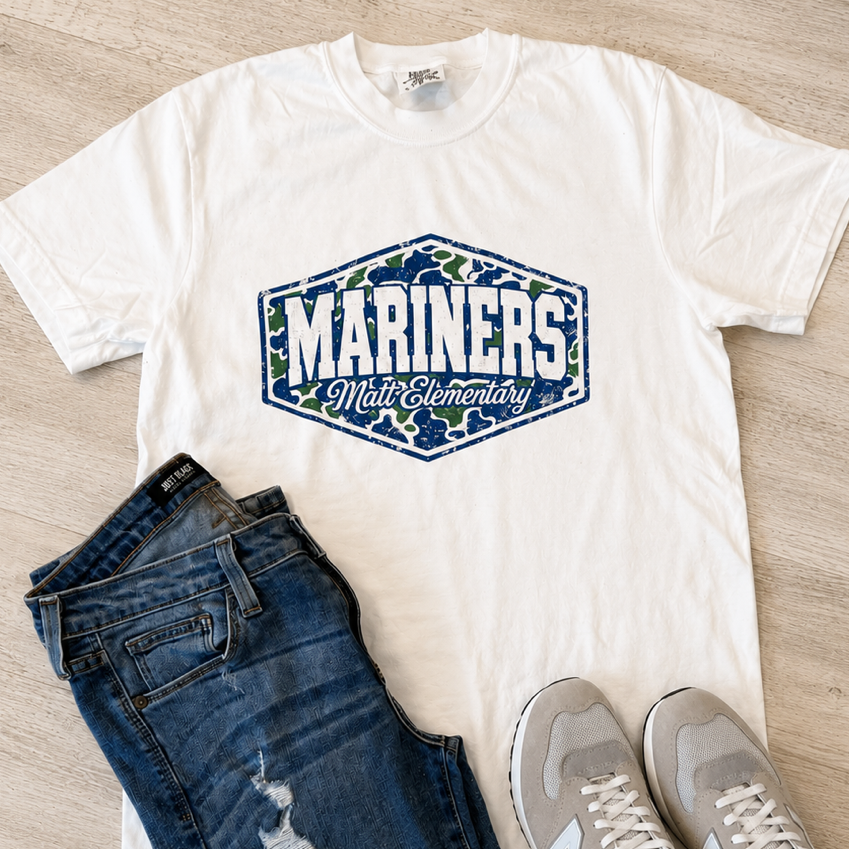 A white graphic T-shirt with a blue and green 'Mariners Who Care' design, styled in a flat lay with dark blue jeans and gray New Balance sneakers on a light wood floor.