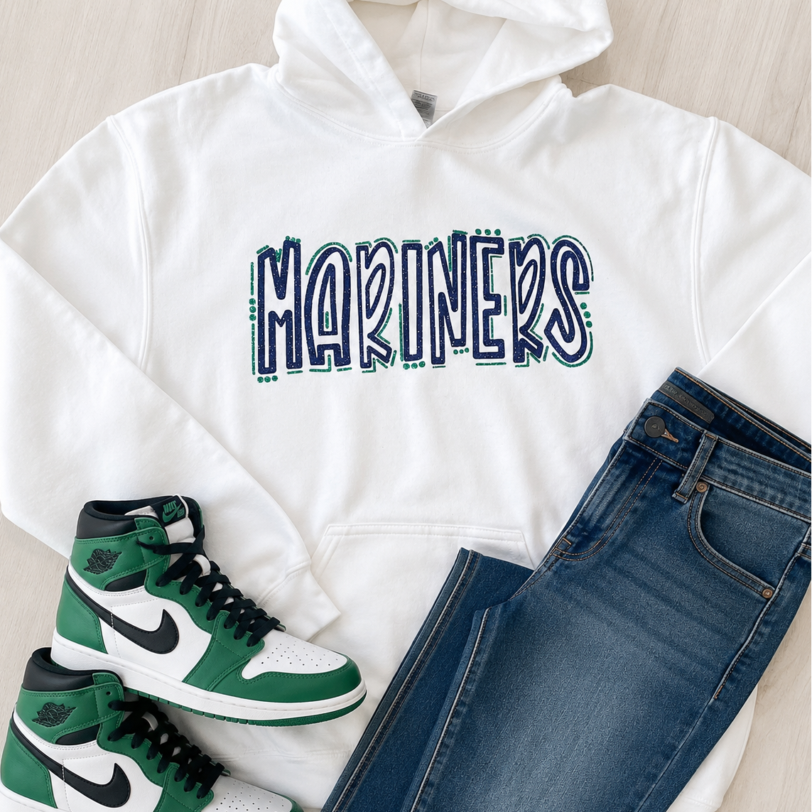 A styled flat-lay outfit featuring a white hoodie with 'MARINERS' printed across the chest in navy and green, paired with blue jeans and green-and-white sneakers on a light wood background.