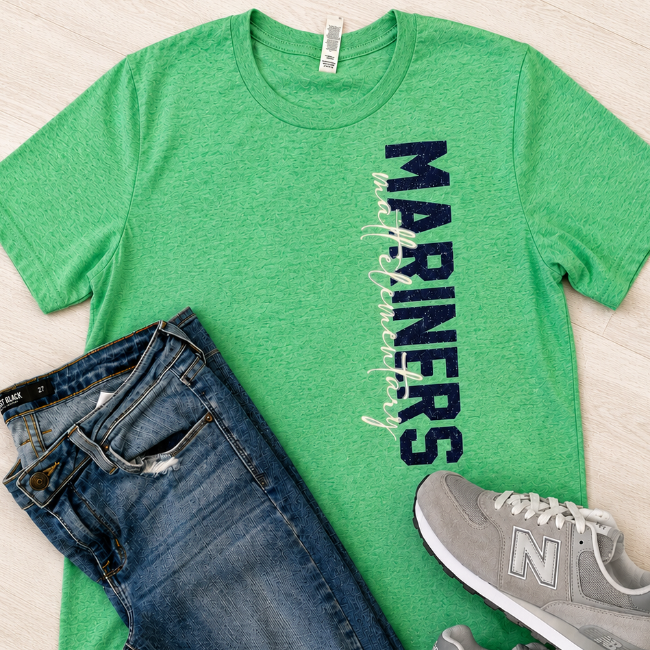 A green heather crewneck t-shirt with 'MARINERS' printed vertically in dark blue, styled in a flat lay with blue jeans and gray New Balance sneakers on a light wood floor.