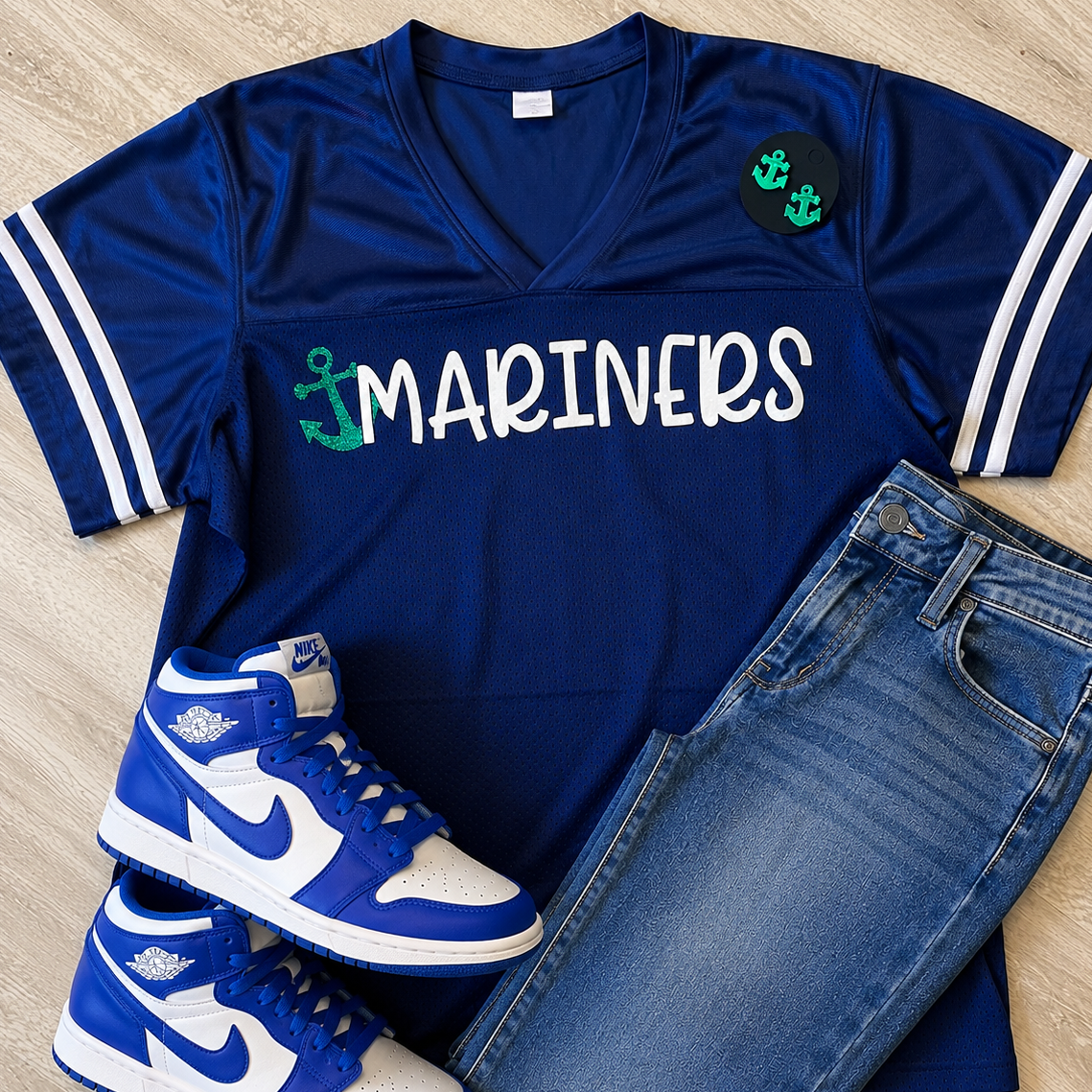 A flat-lay outfit arrangement featuring a royal blue V-neck athletic jersey with 'MARINERS' in white and green anchor graphics, paired with blue jeans and blue-and-white sneakers on a light wood floor.