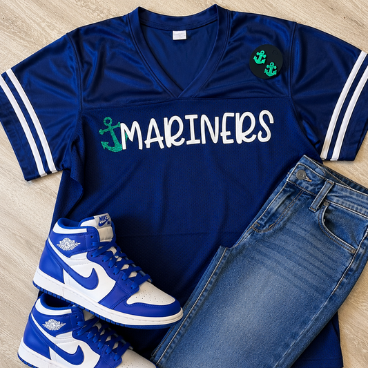 A flat-lay outfit arrangement featuring a royal blue V-neck athletic jersey with 'MARINERS' in white and green anchor graphics, paired with blue jeans and blue-and-white sneakers on a light wood floor.
