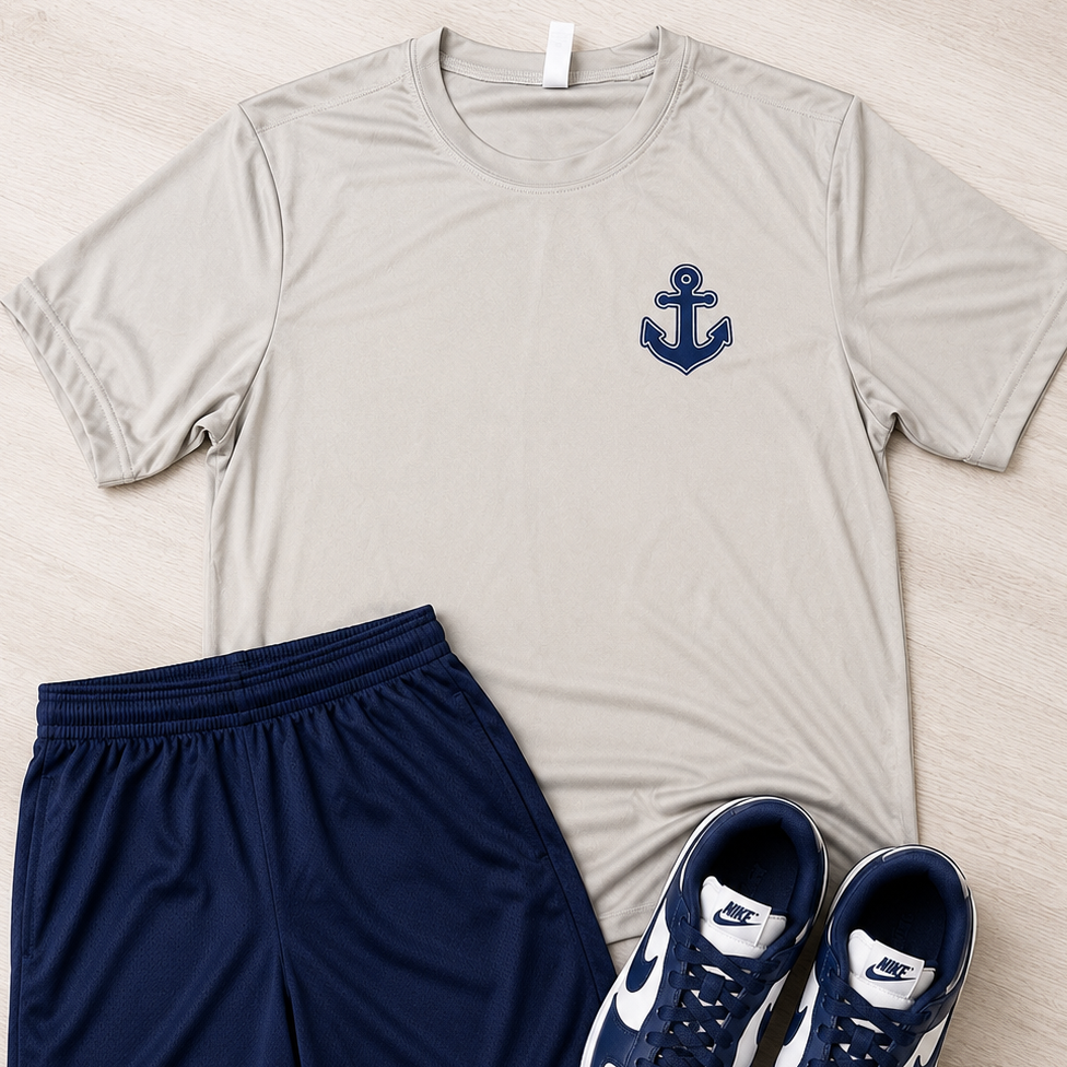 A flat-lay outfit set featuring a light beige short-sleeve athletic t-shirt with a navy blue anchor logo on the chest, paired with navy blue athletic shorts and navy and white Nike sneakers, all arranged on a light wood background.