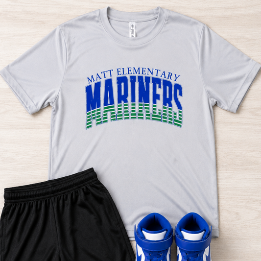 A flat-lay outfit set featuring a light gray t-shirt with a blue and green 'Matt Elementary Mariners' graphic, black athletic shorts, and blue and white sneakers, all arranged on a light wood background.