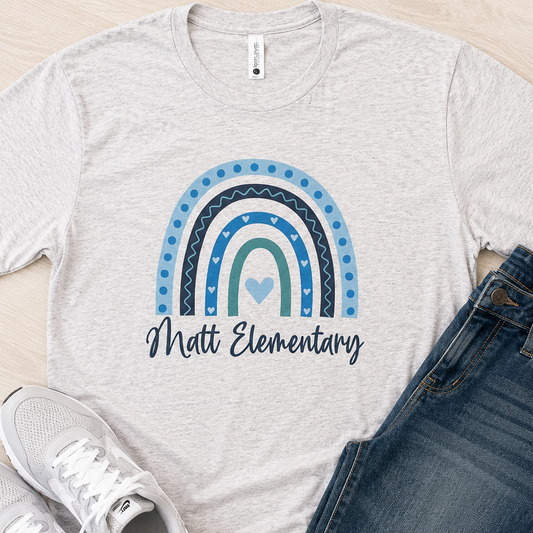 A light heather gray t-shirt with a blue and teal rainbow graphic and the text 'Matt Elementary' printed on the front, styled in a flat lay with blue jeans and white sneakers on a light wood floor.