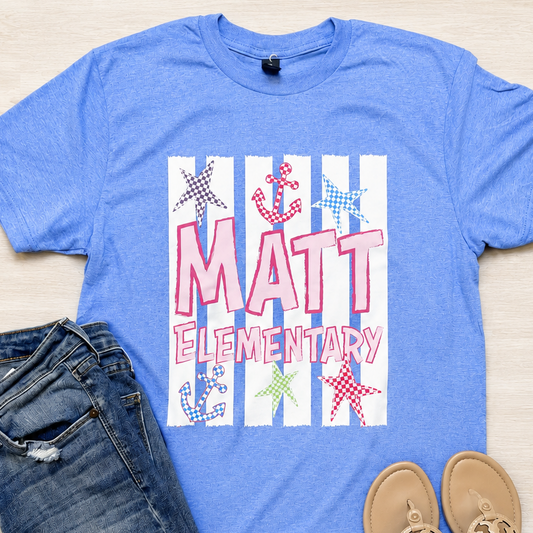 A blue graphic t-shirt with 'MATT ELEMENTARY' and nautical designs, styled in a flat lay with blue jeans and tan slide sandals on a light wood background.
