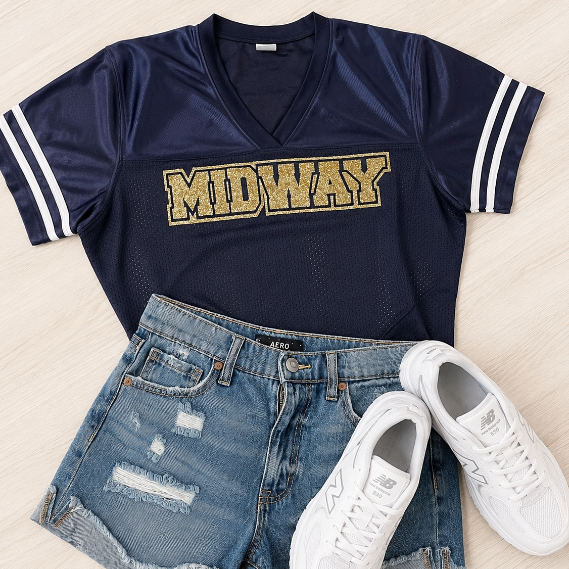 A navy blue athletic-style V-neck T-shirt with white sleeve stripes and a glittery gold 'MIDWAY' graphic across the chest, styled in a flat lay with distressed denim shorts and white sneakers on a light wood background.