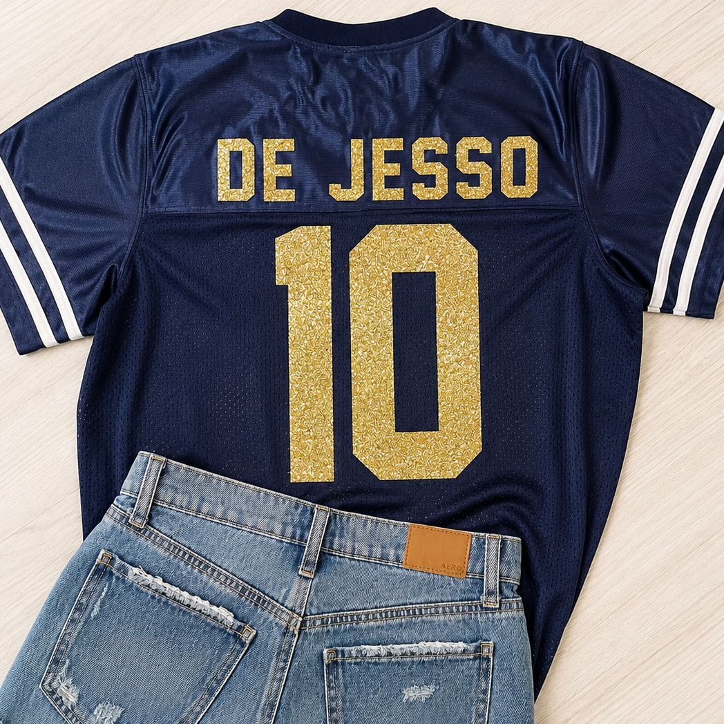 A navy blue football-style jersey with 'DE JESSO' and the number '10' in glittery gold on the back, styled flat-lay with light-wash denim shorts on a light wood background.