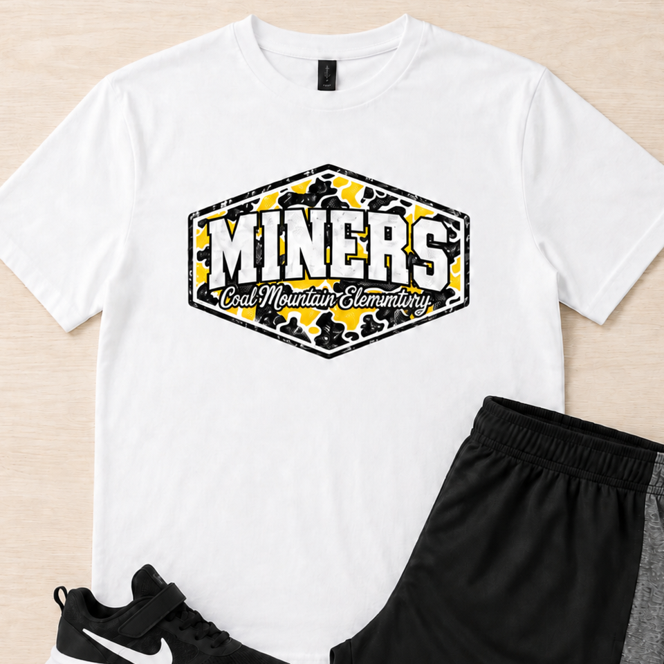 A flat-lay outfit set featuring a white t-shirt with a 'MINERS Cal Mountain Seminary' graphic, black athletic shorts with a gray side panel, and a pair of black Nike sneakers, arranged on a light wood background.