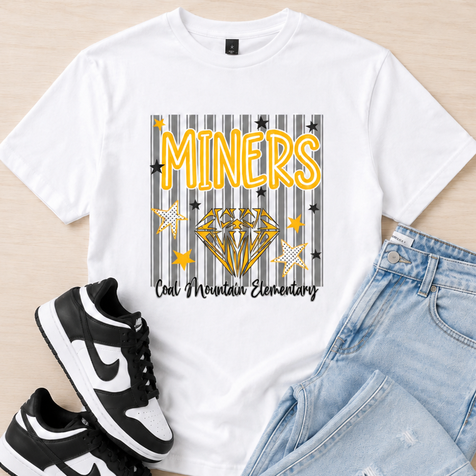 A white graphic t-shirt featuring a 'Miners' design with a diamond and school name, styled in a flat lay with light blue jeans and black-and-white sneakers on a light wood background.
