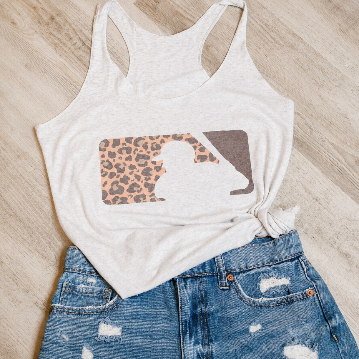 A light heather gray racerback tank top with a graphic print of a silhouette inside a leopard-print and solid brown shape, styled on a light wood floor with blue distressed denim shorts.