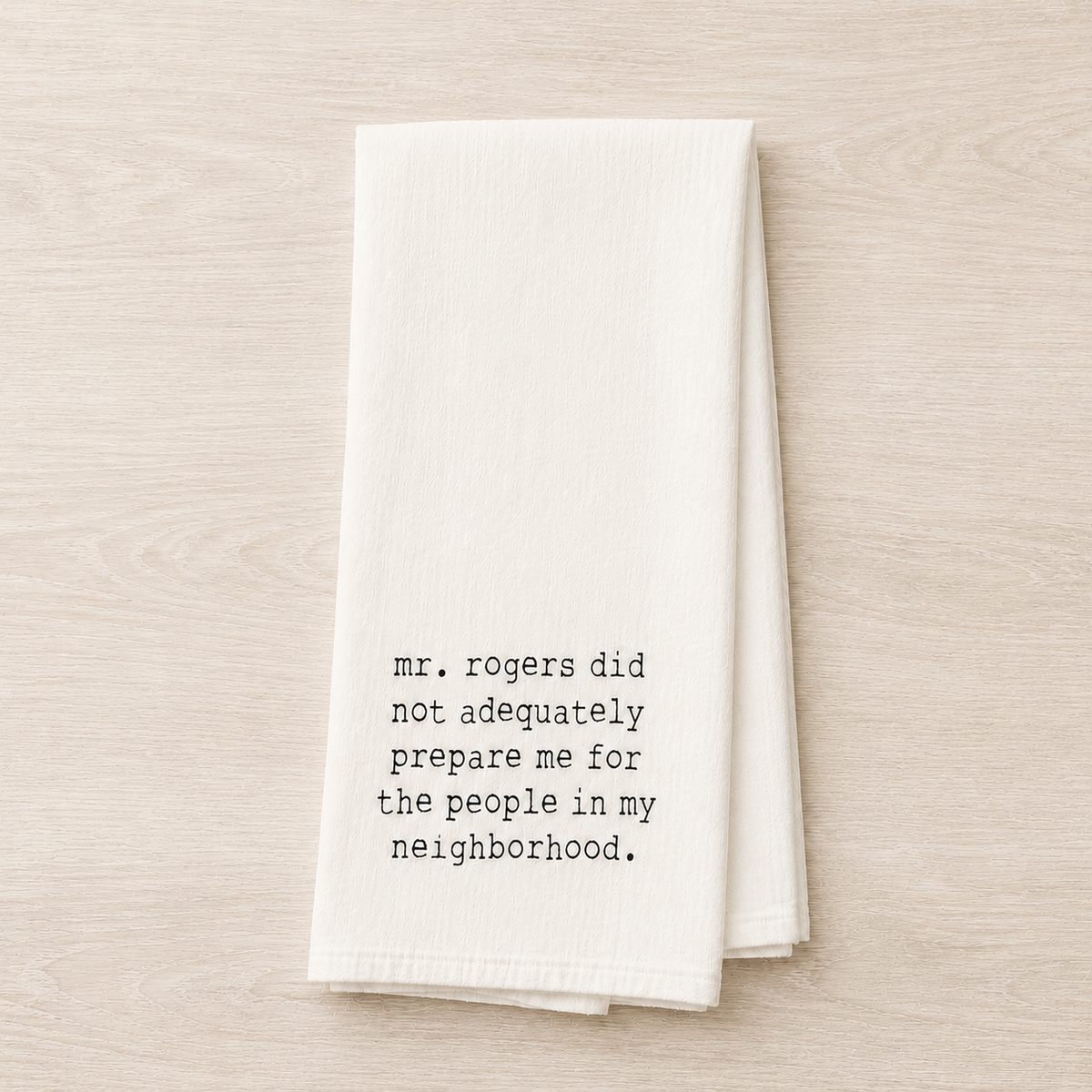 A folded white kitchen towel with black printed text reading 'mr. rogers did not adequately prepare me for the people in my neighborhood.
